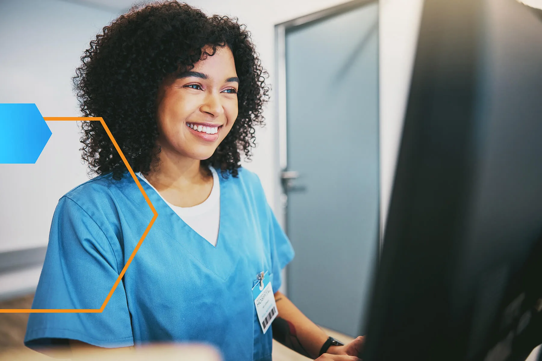 Smiling healthcare worker in blue scrubs using a computer at a medical facility, wearing a name badge and seated in a brightly lit room.