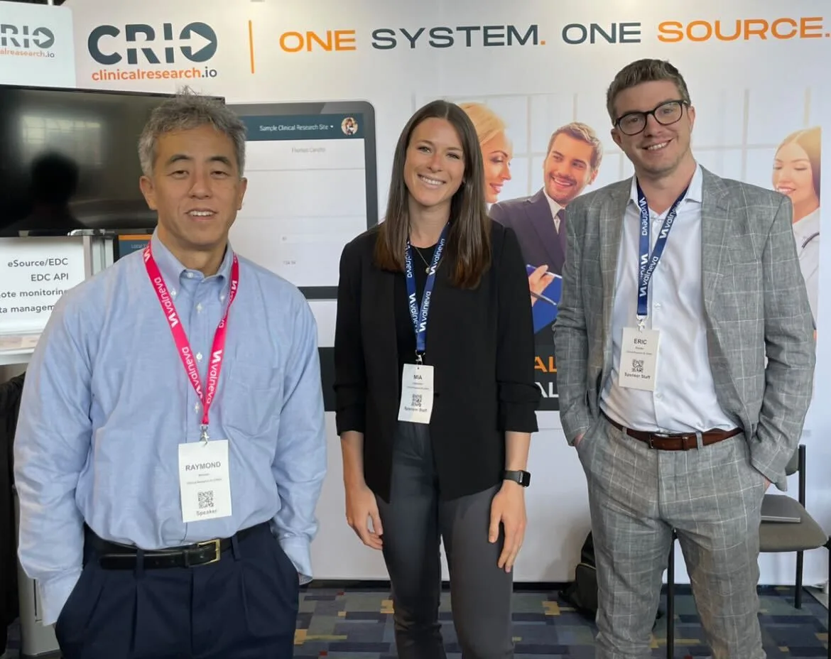 Three professionals standing in front of a CRIO clinical research booth at a conference, wearing name badges and smiling at the camera.