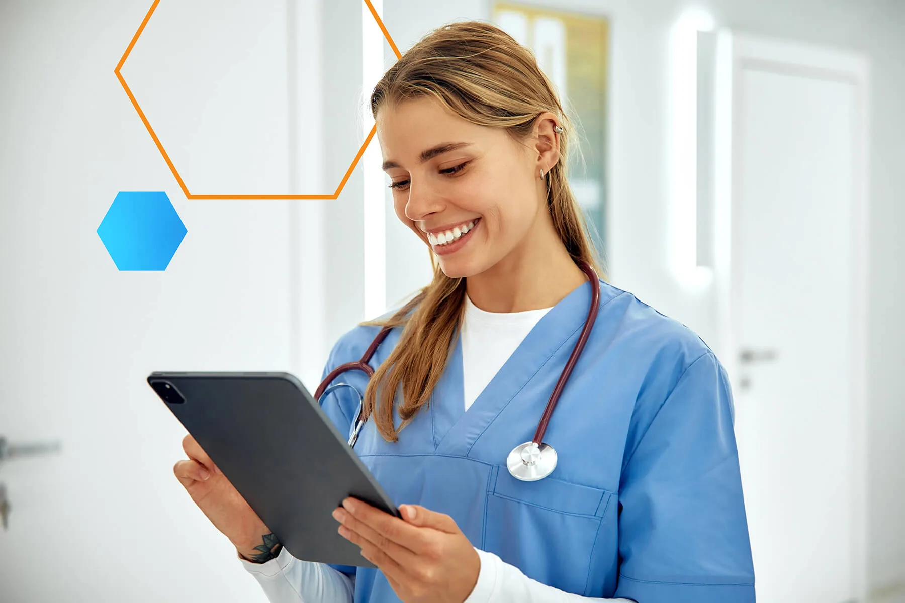 Smiling female healthcare professional in blue scrubs holding a tablet and standing in a bright medical facility, with a stethoscope around her neck.