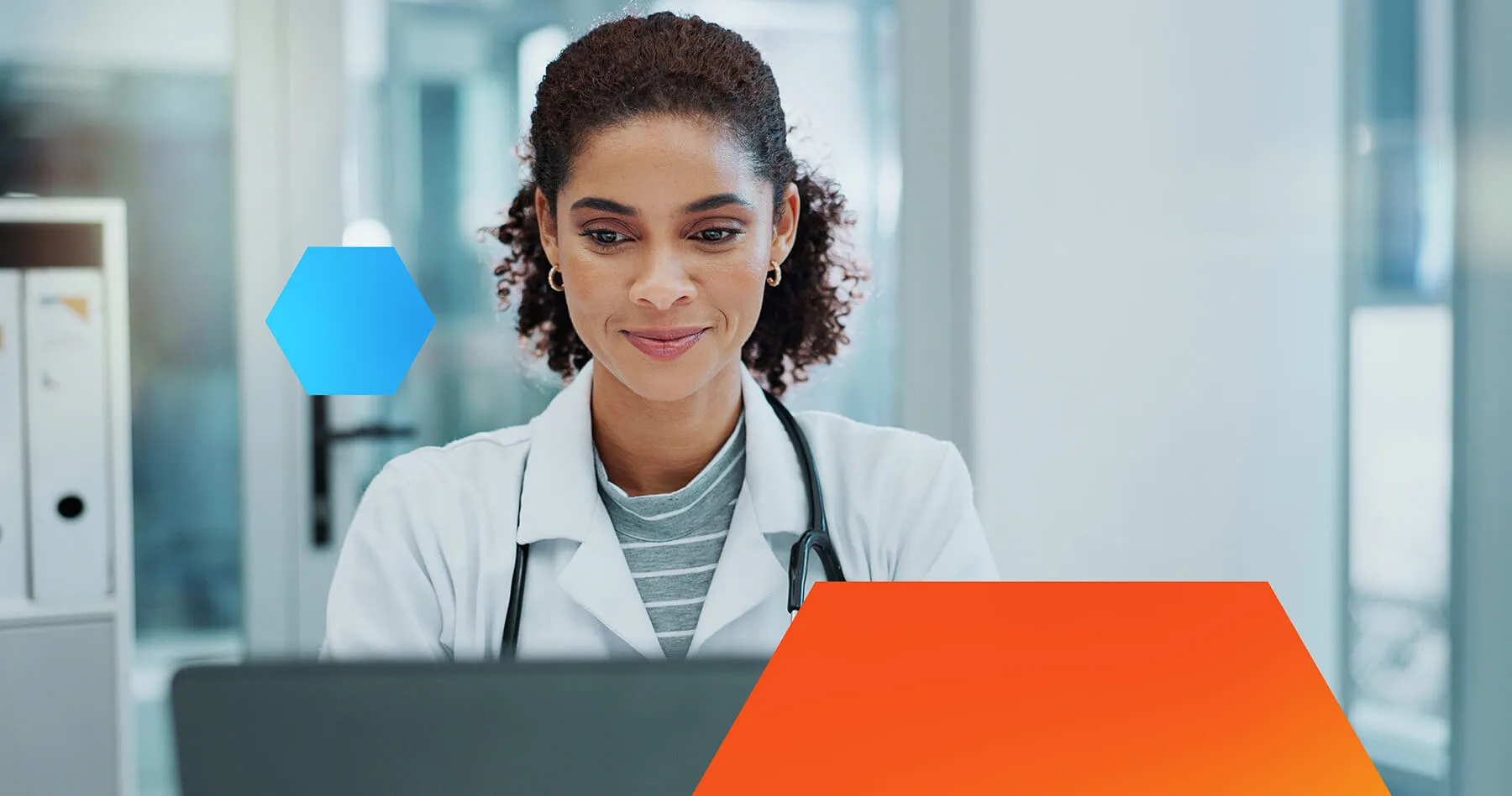 Female doctor with a stethoscope around her neck looking at a laptop screen in a bright medical office, appearing focused and content.
