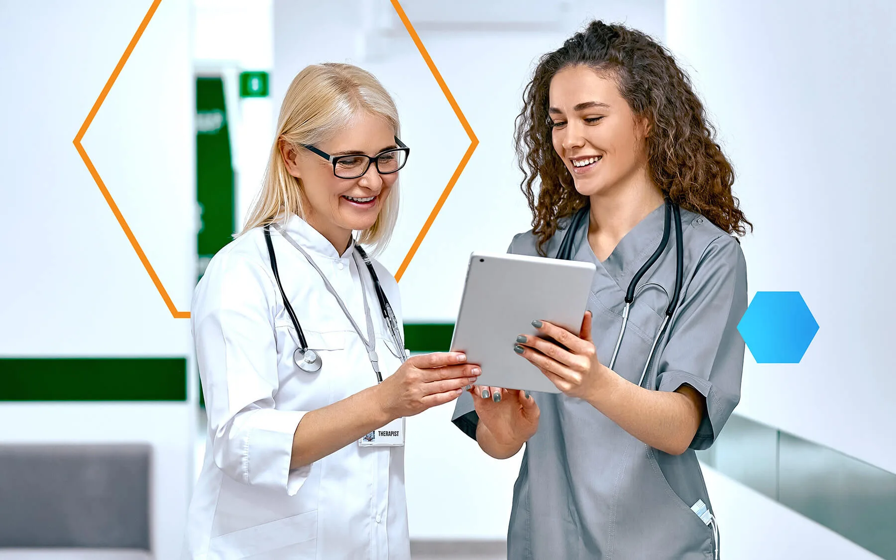 Two female doctors smiling and discussing information on a tablet in a bright hospital corridor.