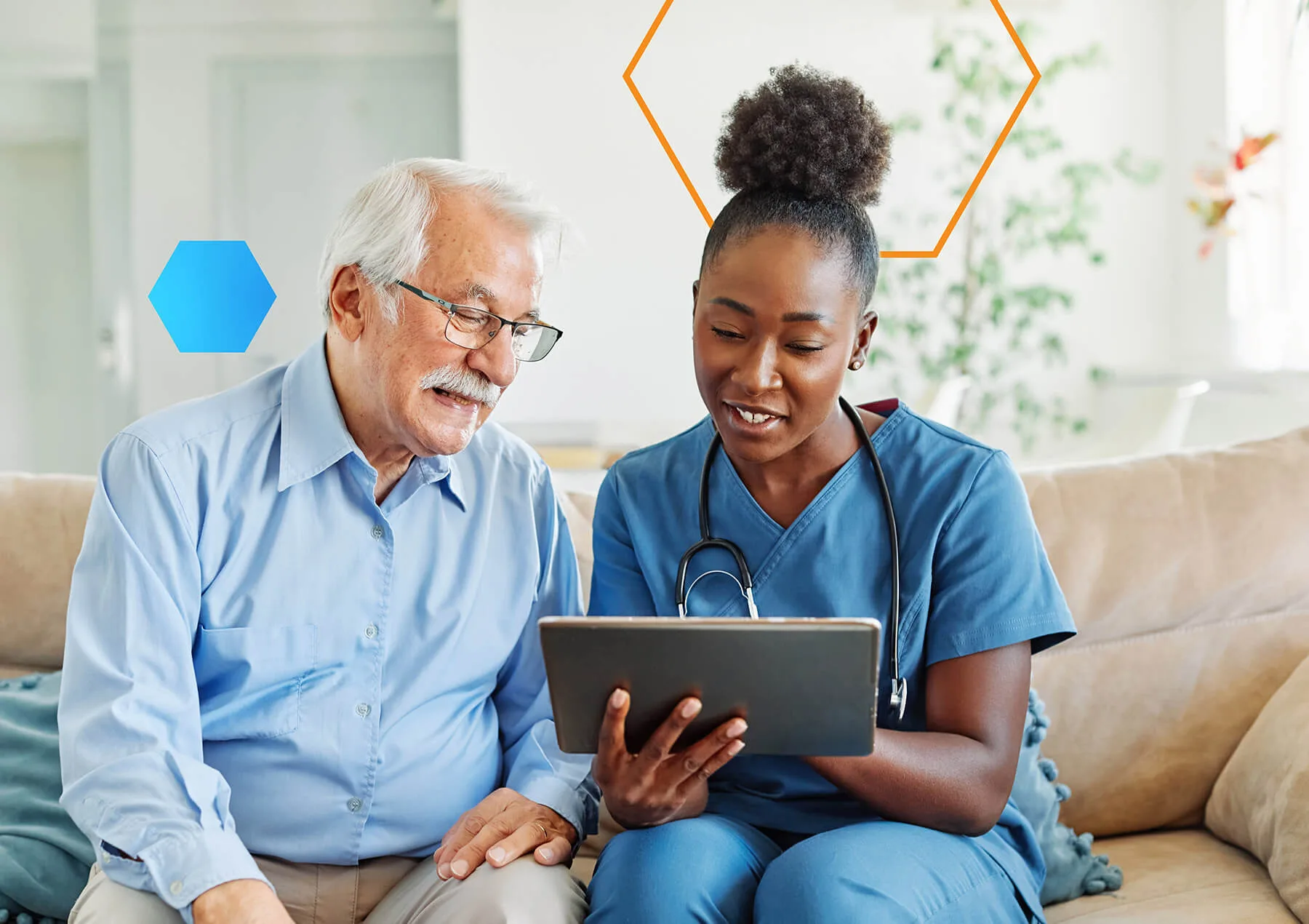 A nurse in blue scrubs showing a tablet to an elderly male patient while sitting together on a sofa.