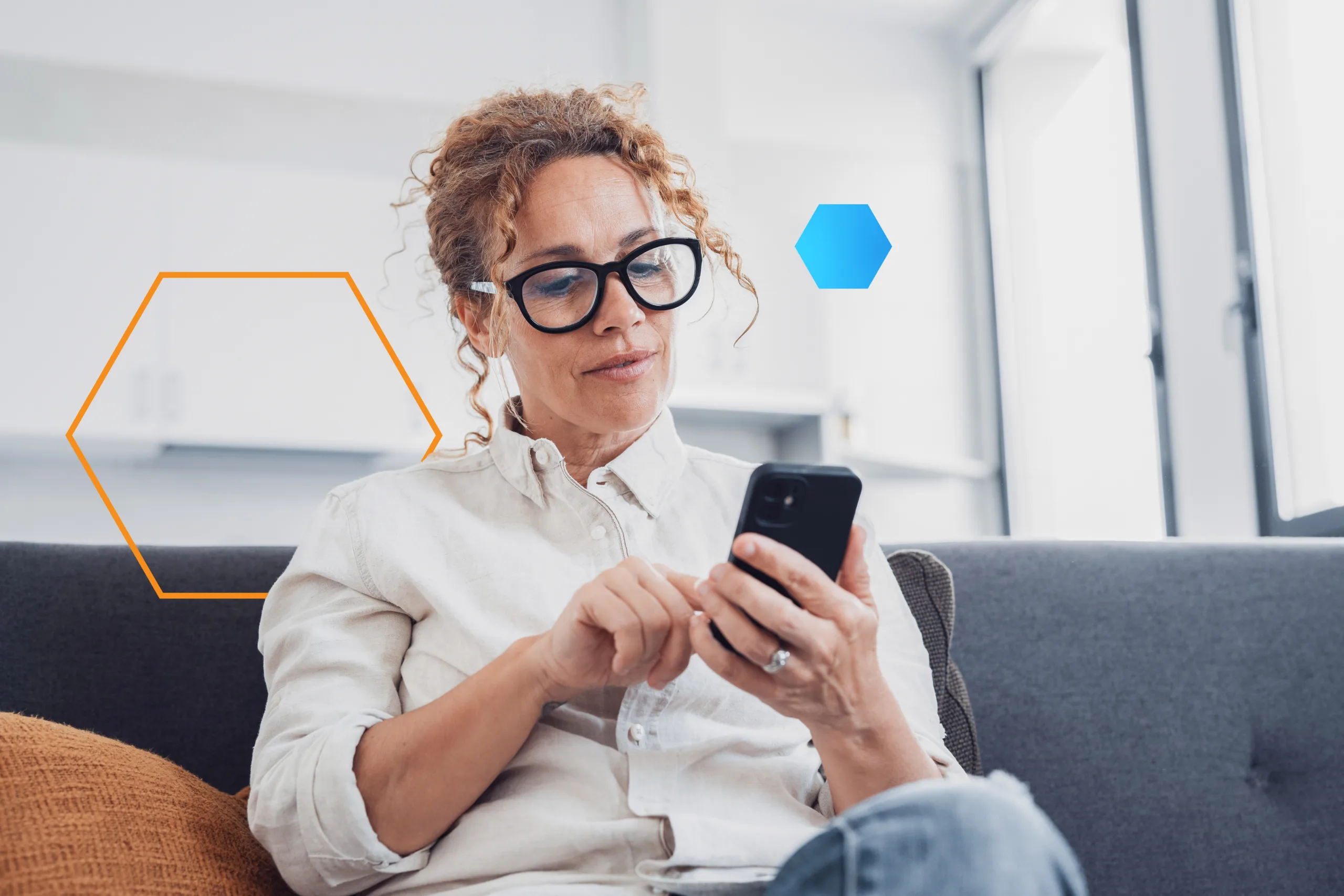 A middle‑aged woman with curly brown hair pulled back, wearing black‑frame glasses and a light button‑up shirt, sits on a gray sofa in a bright, modern living room. She holds a black smartphone in both hands, tapping the screen with her right index finger, and appears focused. Two semi‑transparent hexagon graphics—one orange outline behind her, one solid blue to her right—float in the background.