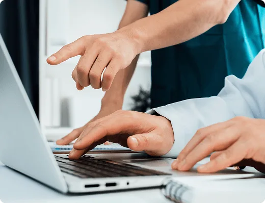 Close-up of two healthcare professionals collaborating at a laptop, with one typing and the other pointing at the screen, indicating teamwork in a clinical or medical setting.