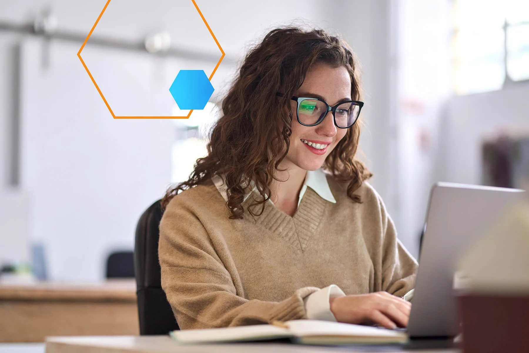 A woman with glasses and curly hair smiling while typing on a laptop in a cozy, modern workspace.